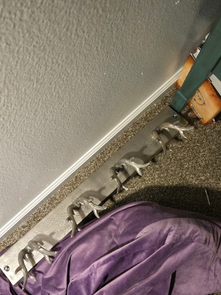 A shadowy photo of a big silver bag holder where the bag hooks are antlers. The holder is on the carpeted floor partially on top my purple meditation mat because it is difficult to mount.