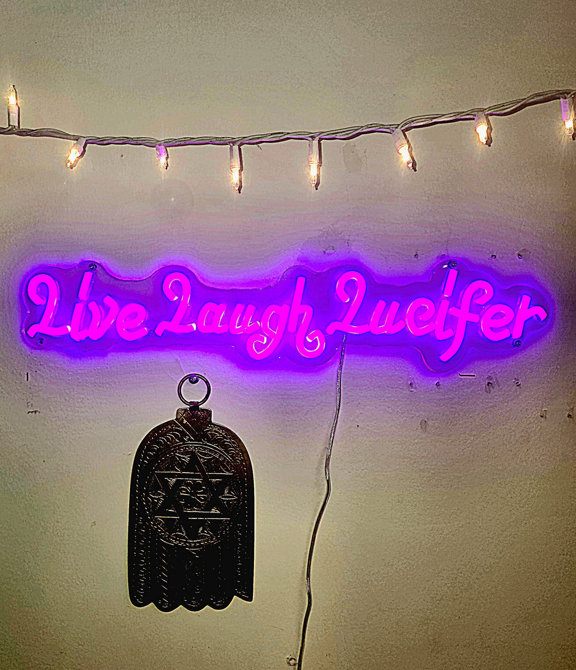 Purple neon sign declares "Live Laugh Lucifer." Hanging right under it by a nail is a metal hand of G-d with a large centered Star of David