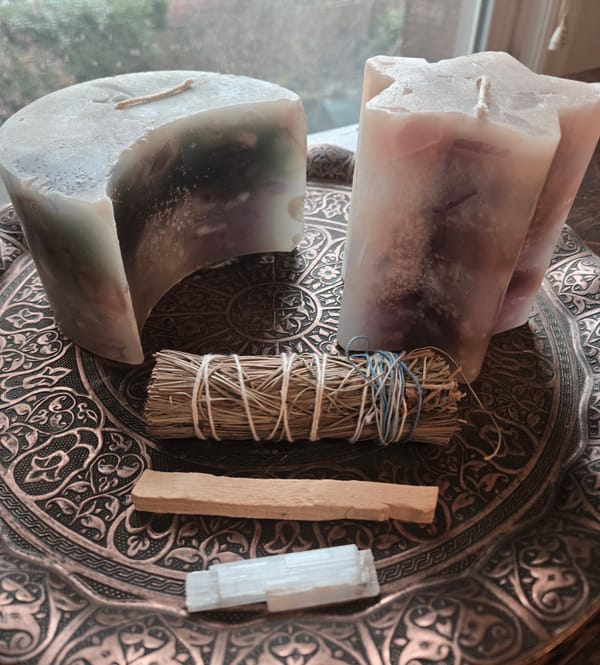 Purple candles shaped like a star and crescent moon on a bronze dish with a pine bundle, palo santo stick, and selenite crystal in front of them